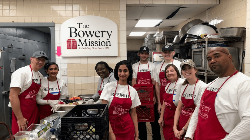 A group of Refinitiv employees gathered in a kitchen with a large banner behind them stating "The Bowery Mission."
