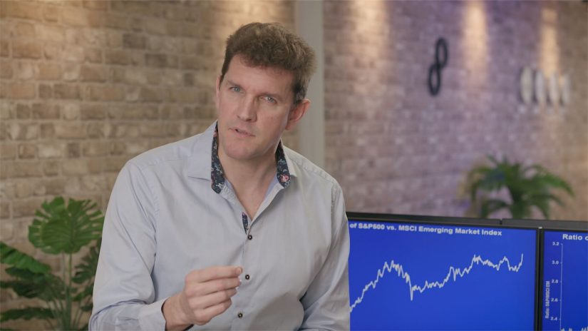Man gestures to camera in an office setting. Two screens behind him display increasing trend charts.